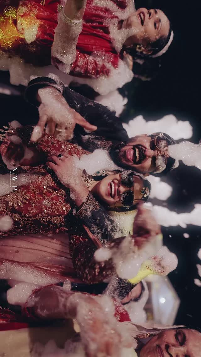 People in traditional Indian wedding attire laughing while playing with white foam bubbles.