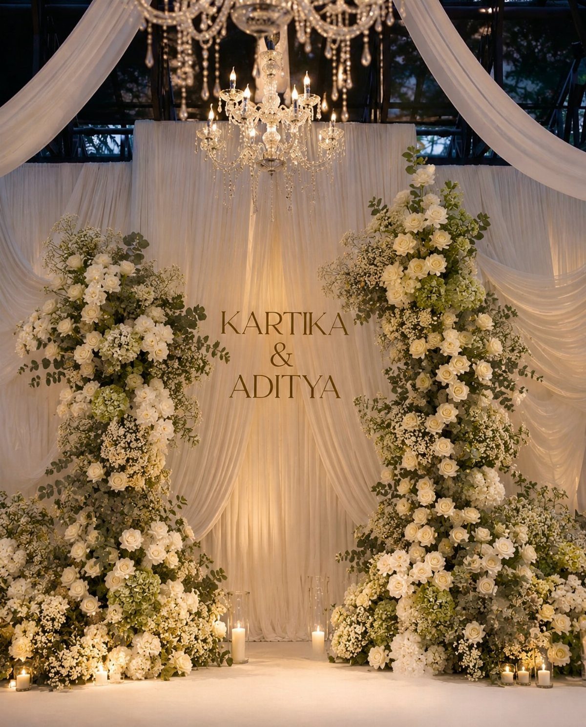 Large white floral arrangements framing a white draped wedding backdrop with chandeliers.