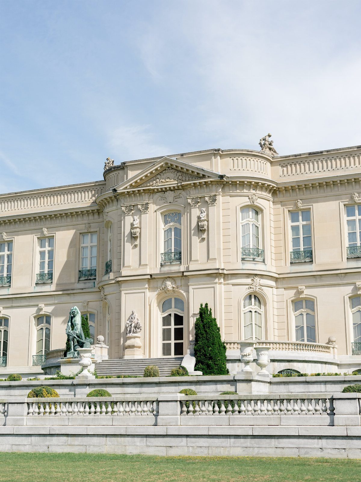 Ornate neoclassical stone mansion with grand staircase and manicured lawn under blue sky