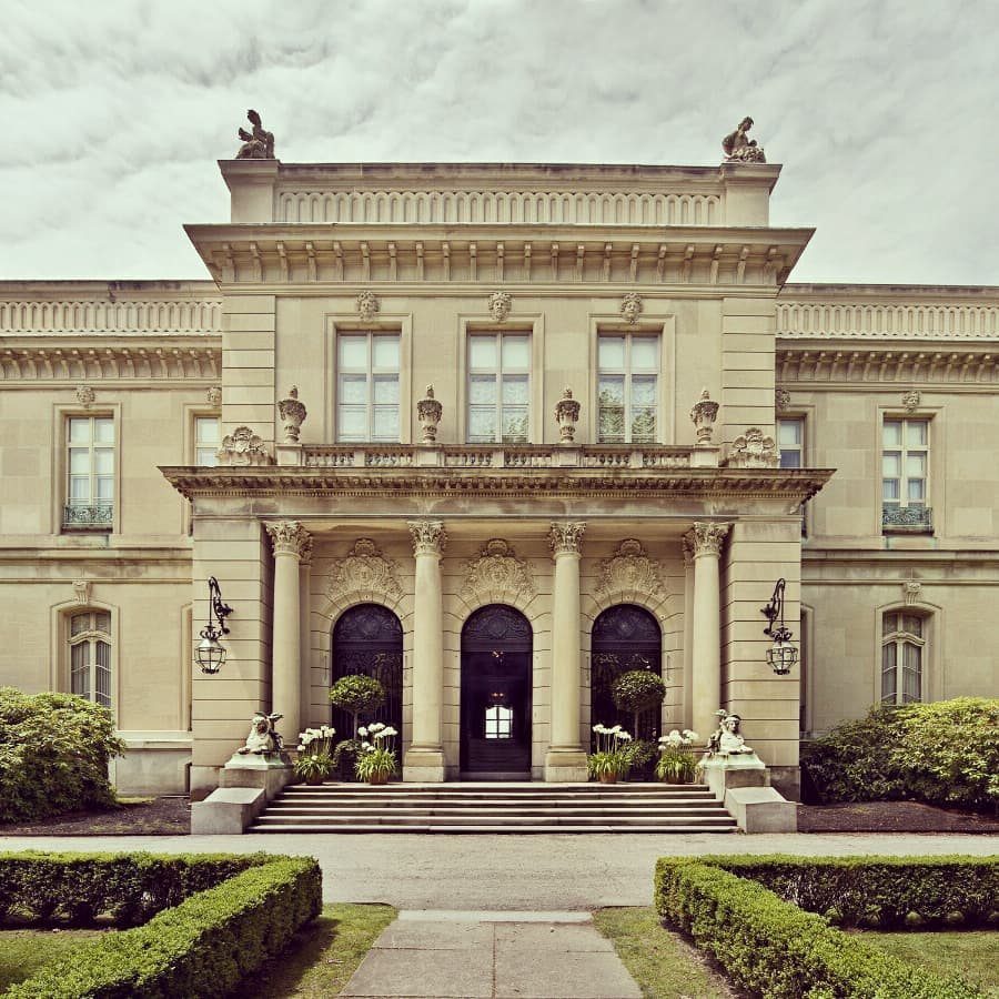 Grand stone mansion with French château architecture, columns, and manicured garden hedges.