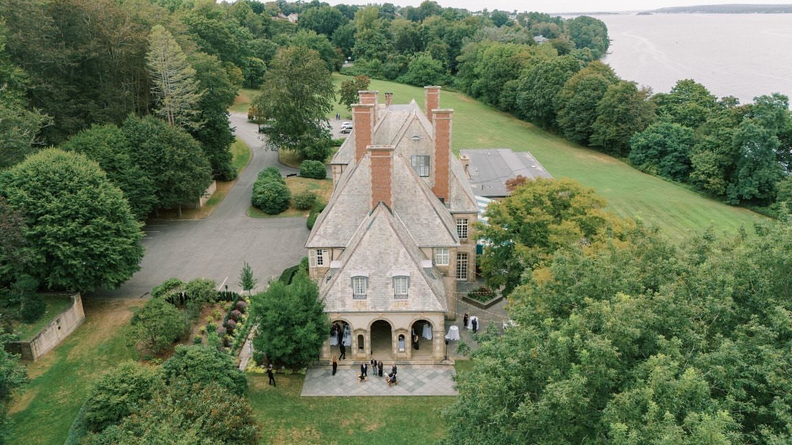 Large stone manor house with steep gables surrounded by green lawns and trees.
