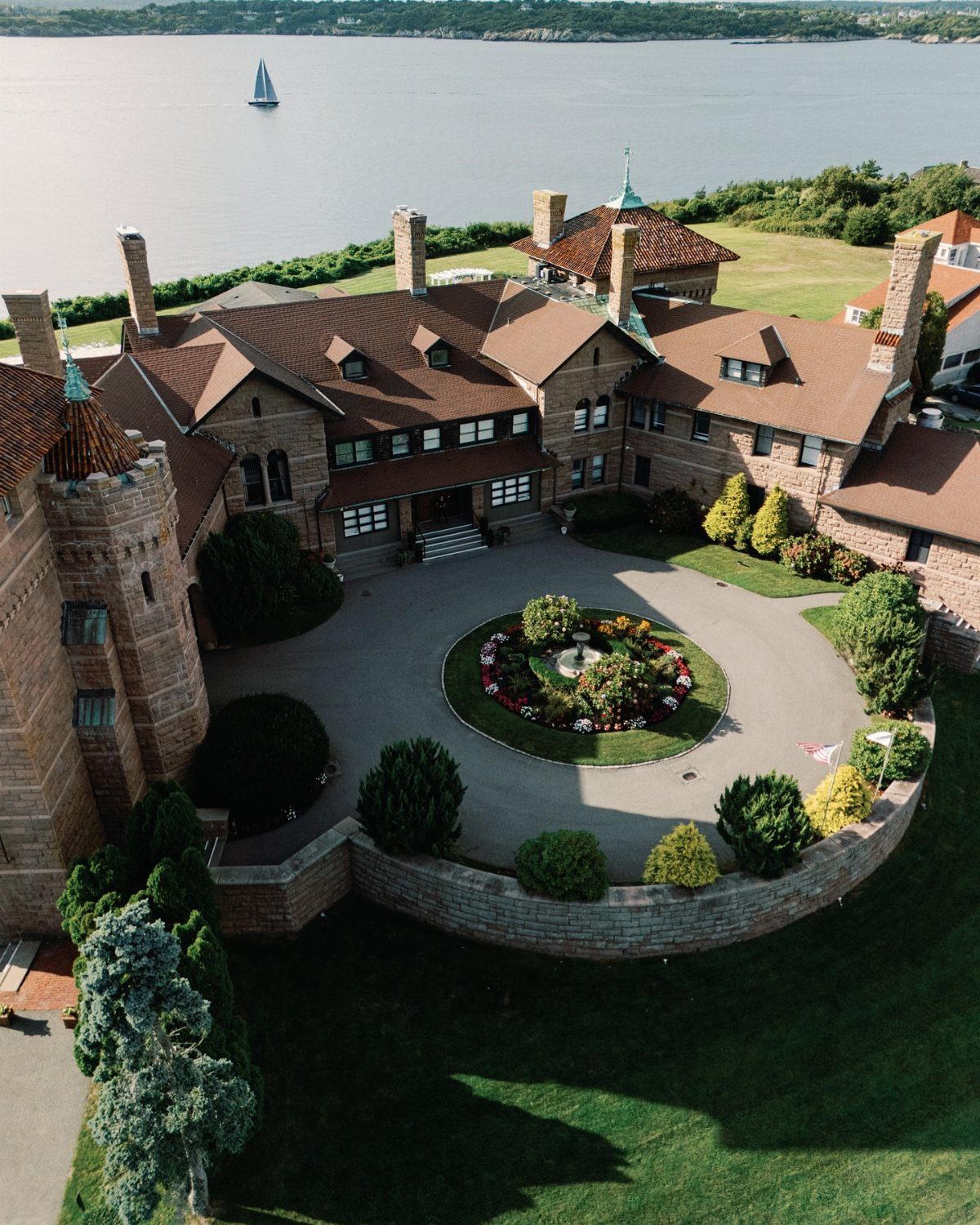 Large stone mansion with brown roofs and circular driveway overlooking a calm bay.