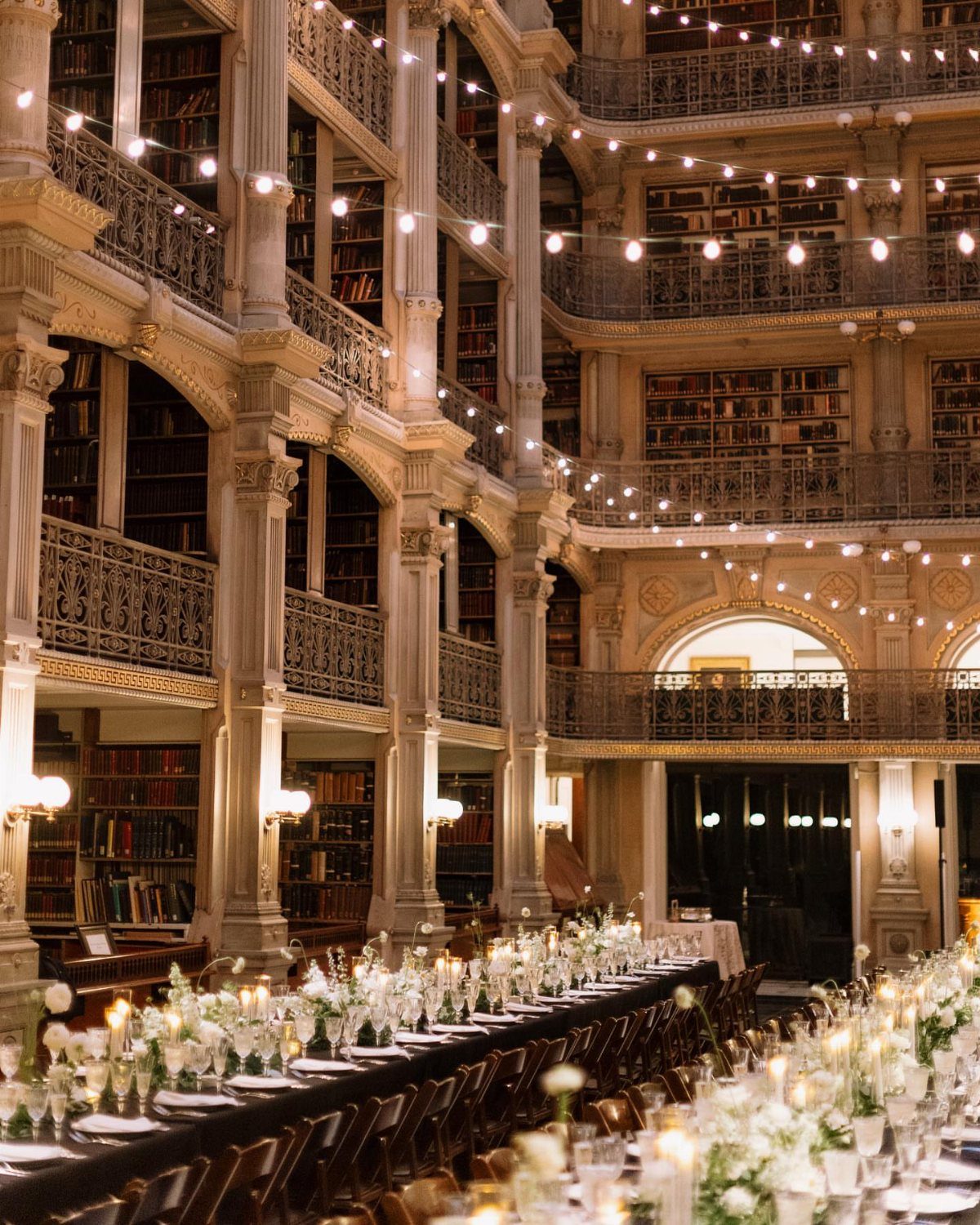 Grand library interior with formal wedding reception tables, white flowers, and warm string lights.