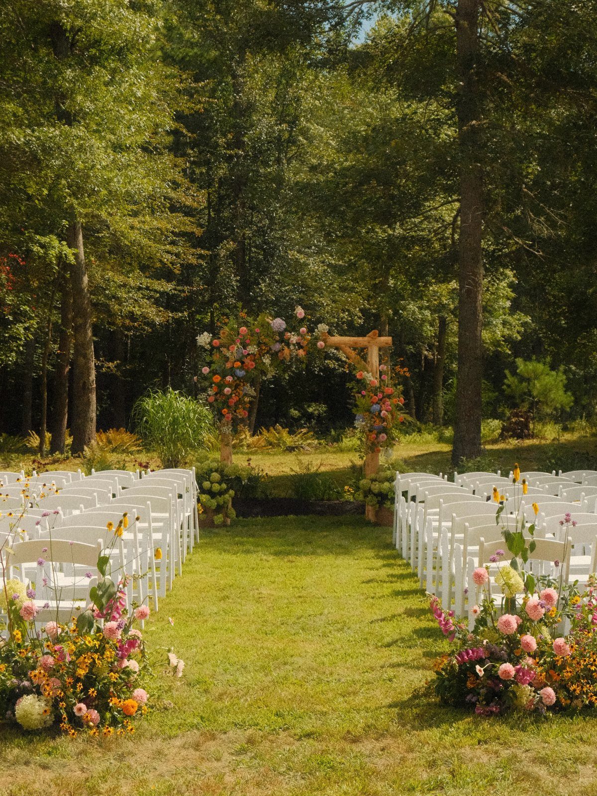Outdoor wedding ceremony setup with white chairs and a floral wooden arch in woods