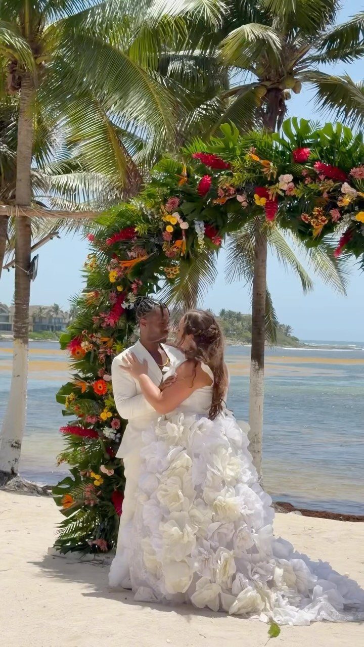 Bride and groom embracing under a floral arch on a tropical sandy beach.