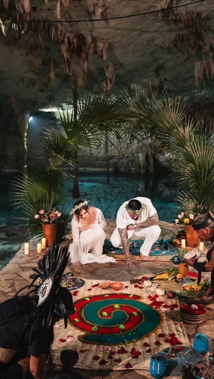Couple in white kneeling during a ceremony inside a limestone cenote with palm arch.