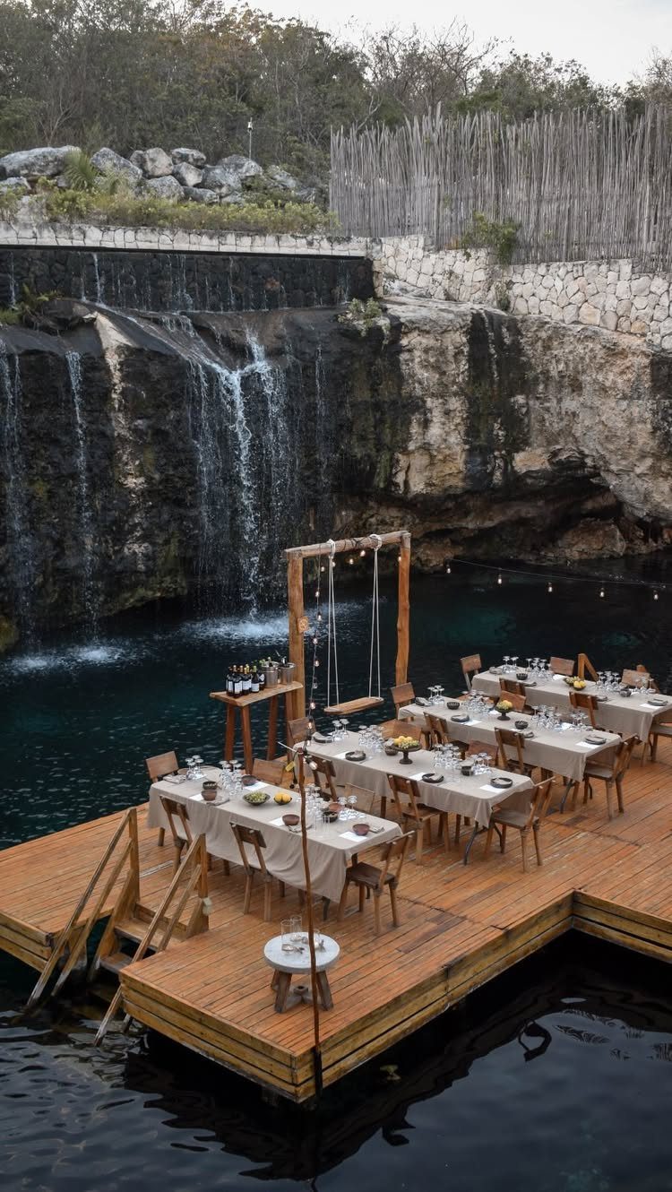 Floating wooden deck with wedding dining tables set before a tropical waterfall backdrop.