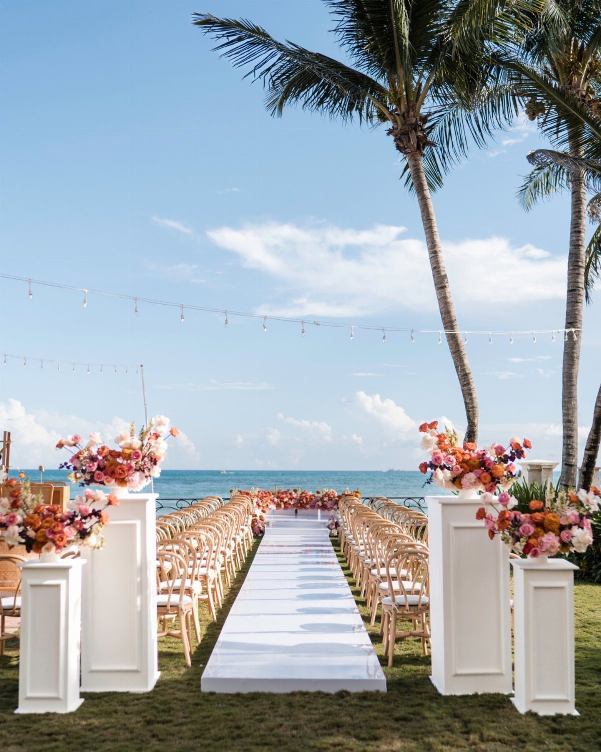 Outdoor wedding ceremony setup with white aisle, gold chairs, and colorful flowers by ocean