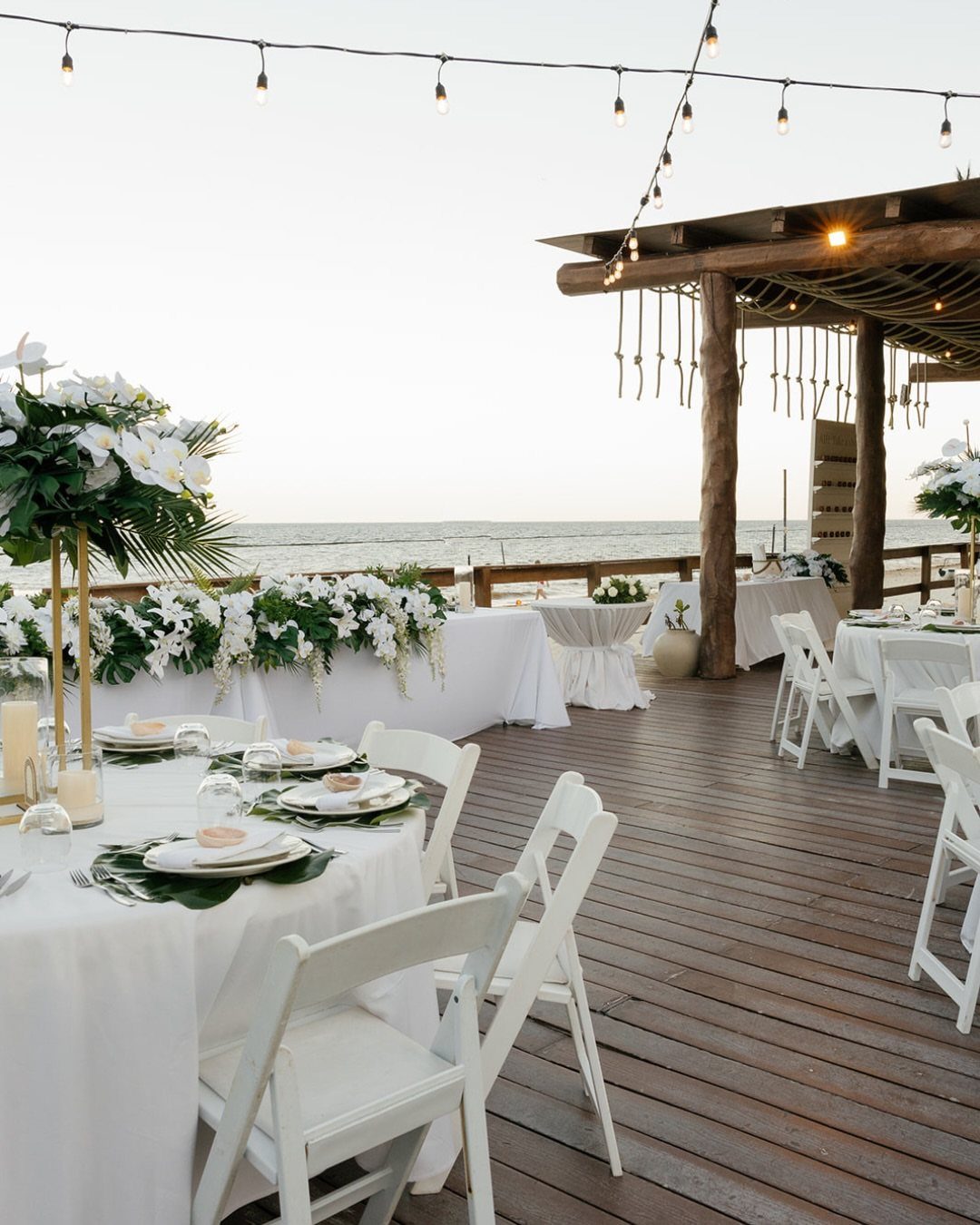 Wedding reception tables with white linens and flowers on a wooden beach deck.