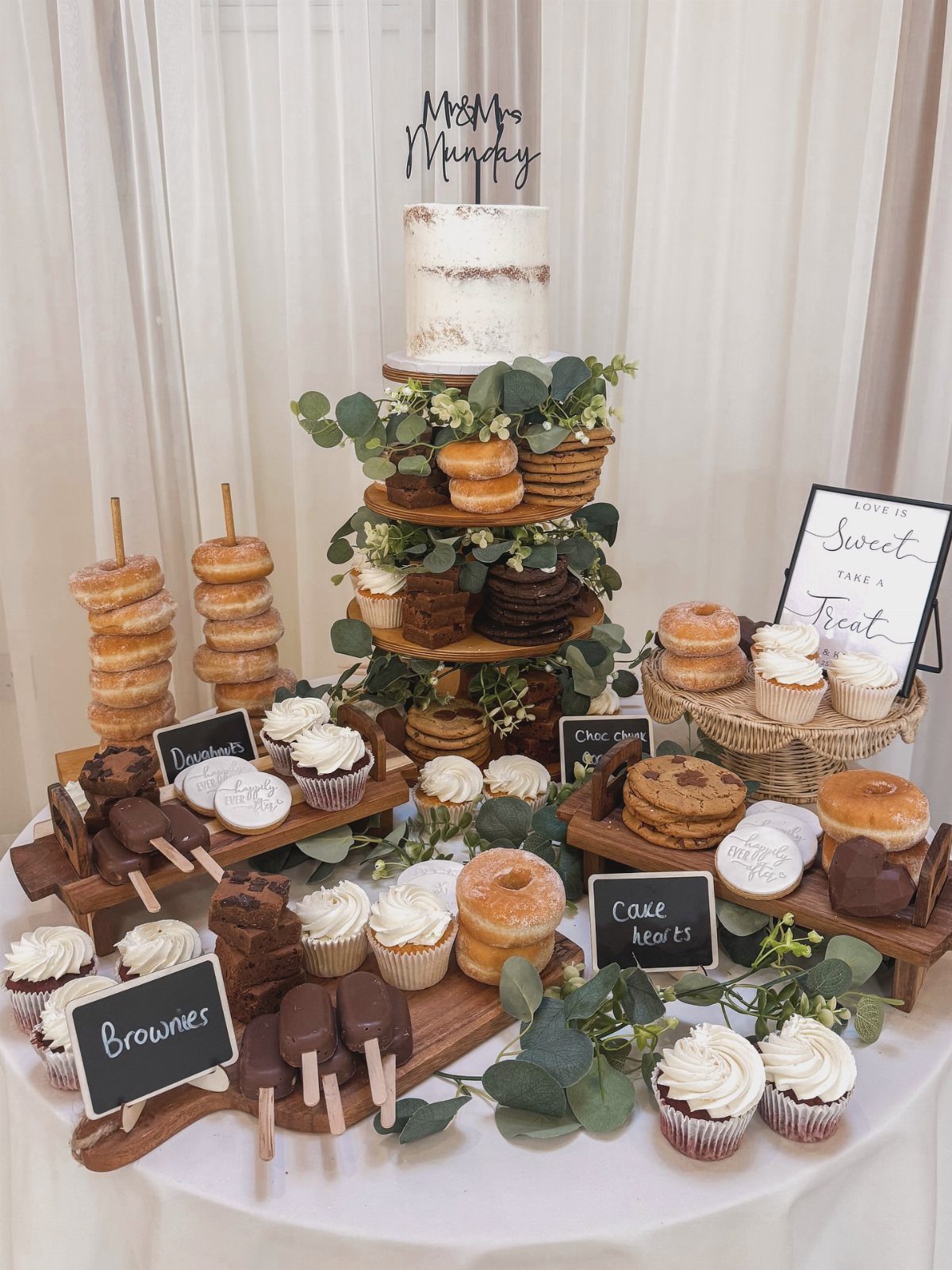 Wedding cake and dessert spread with donuts and cupcakes on rustic wooden stands.