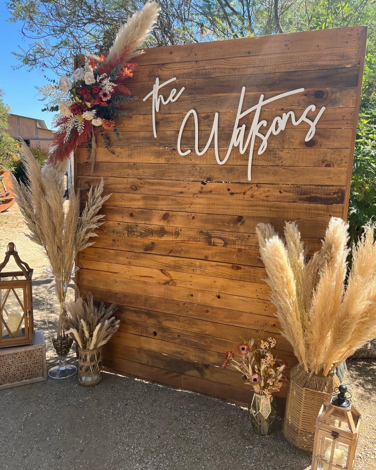 Wooden wall backdrop with white cursive signage, pampas grass, and floral arrangements outdoors.