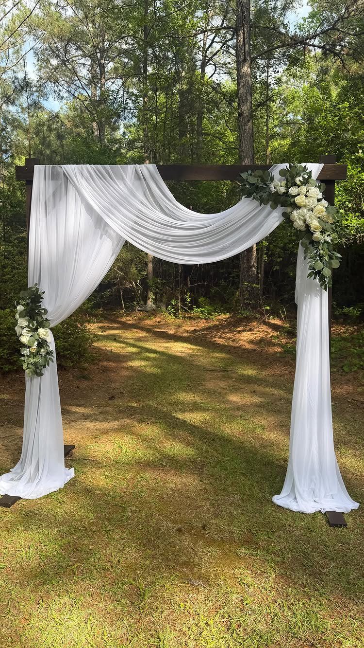 Wooden wedding arch with white fabric draping and white floral arrangements in nature.