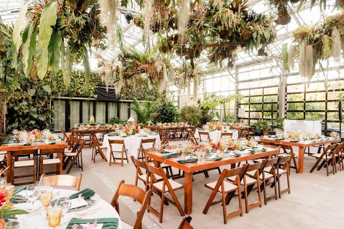 Wedding reception tables set inside a lush glass greenhouse with hanging tropical plants.