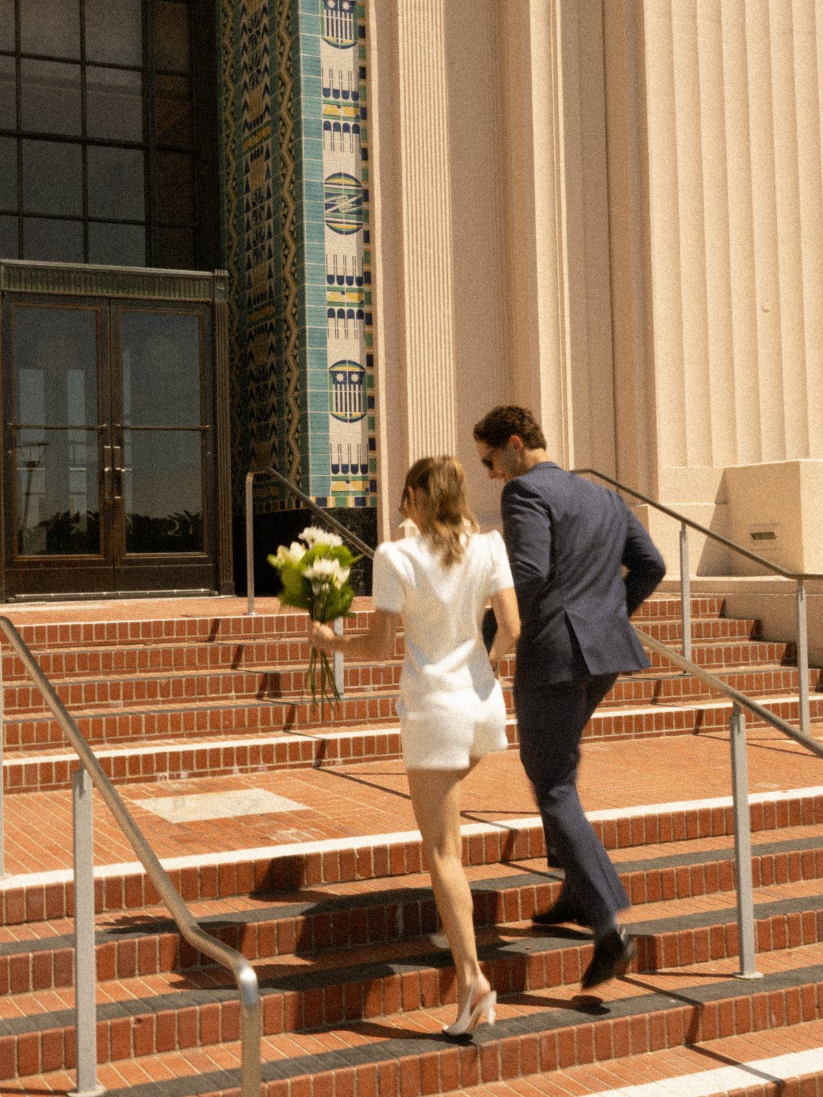 Couple in wedding attire walking up brick steps toward a government building