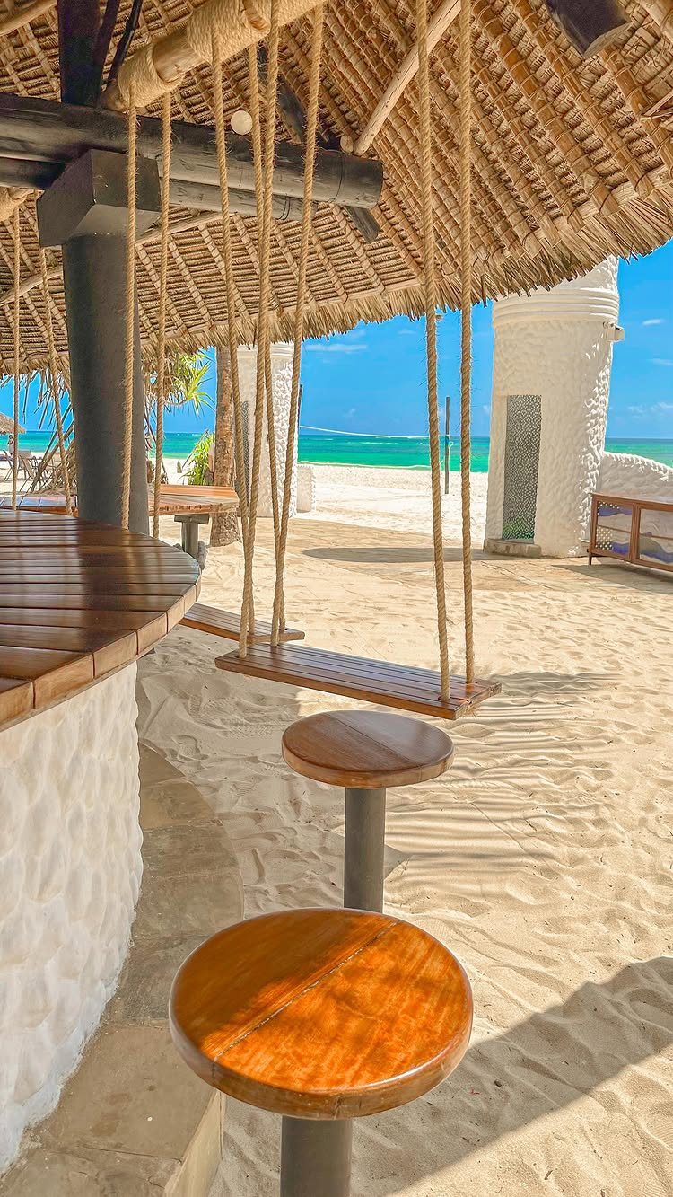 Beach bar with thatched roof, wooden stools, and rope swing on white sand.