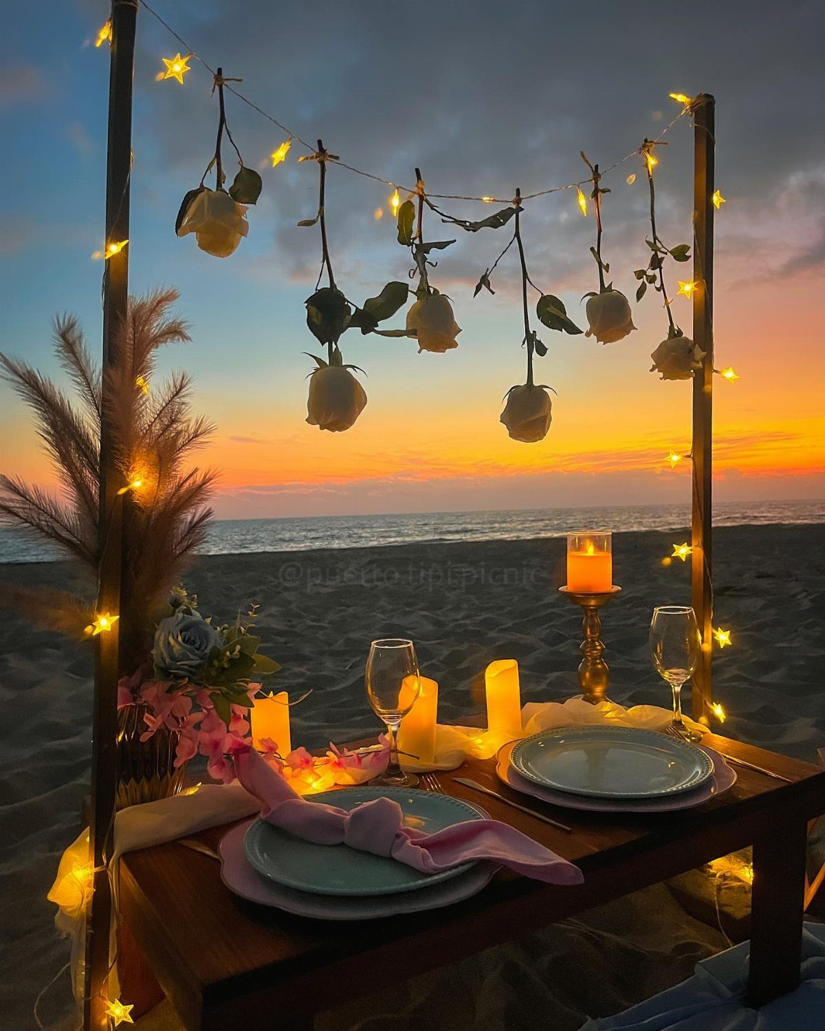 Romantic beach dinner table with white roses and fairy lights at sunset