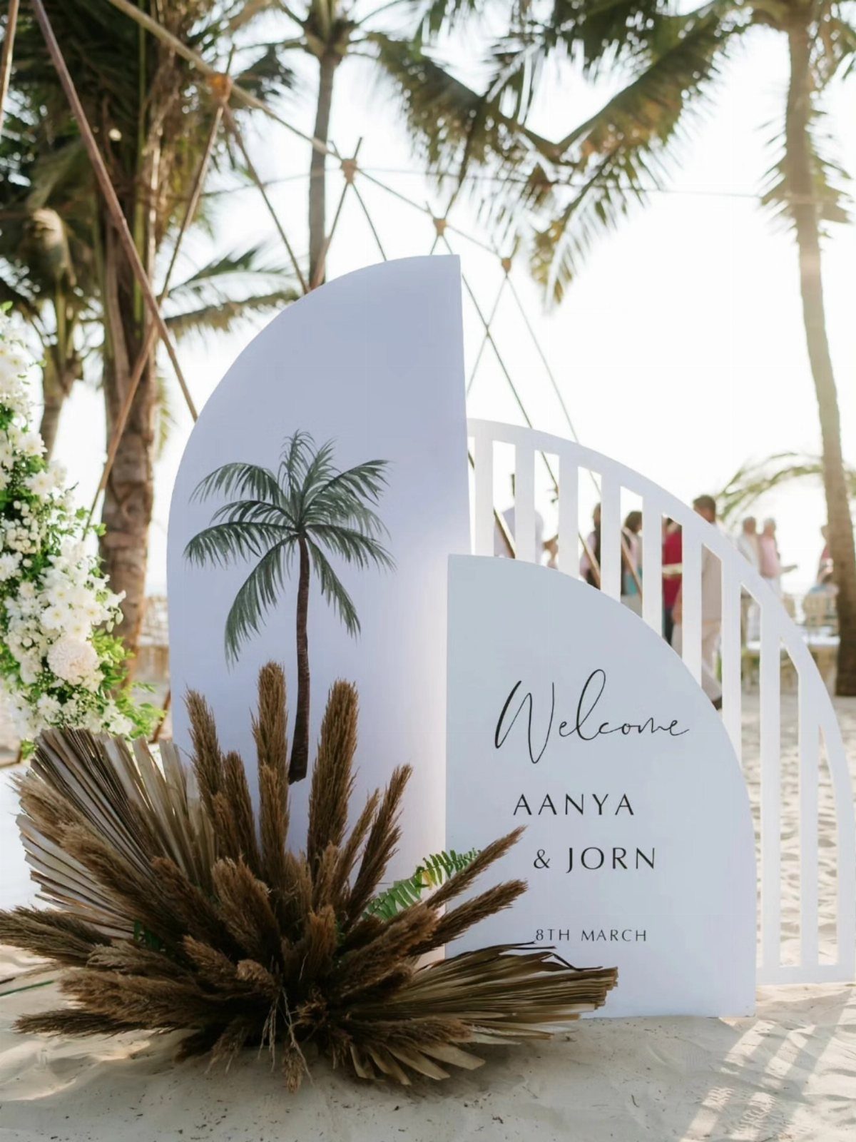 White curved wedding welcome sign with palm tree illustration on a sandy beach.