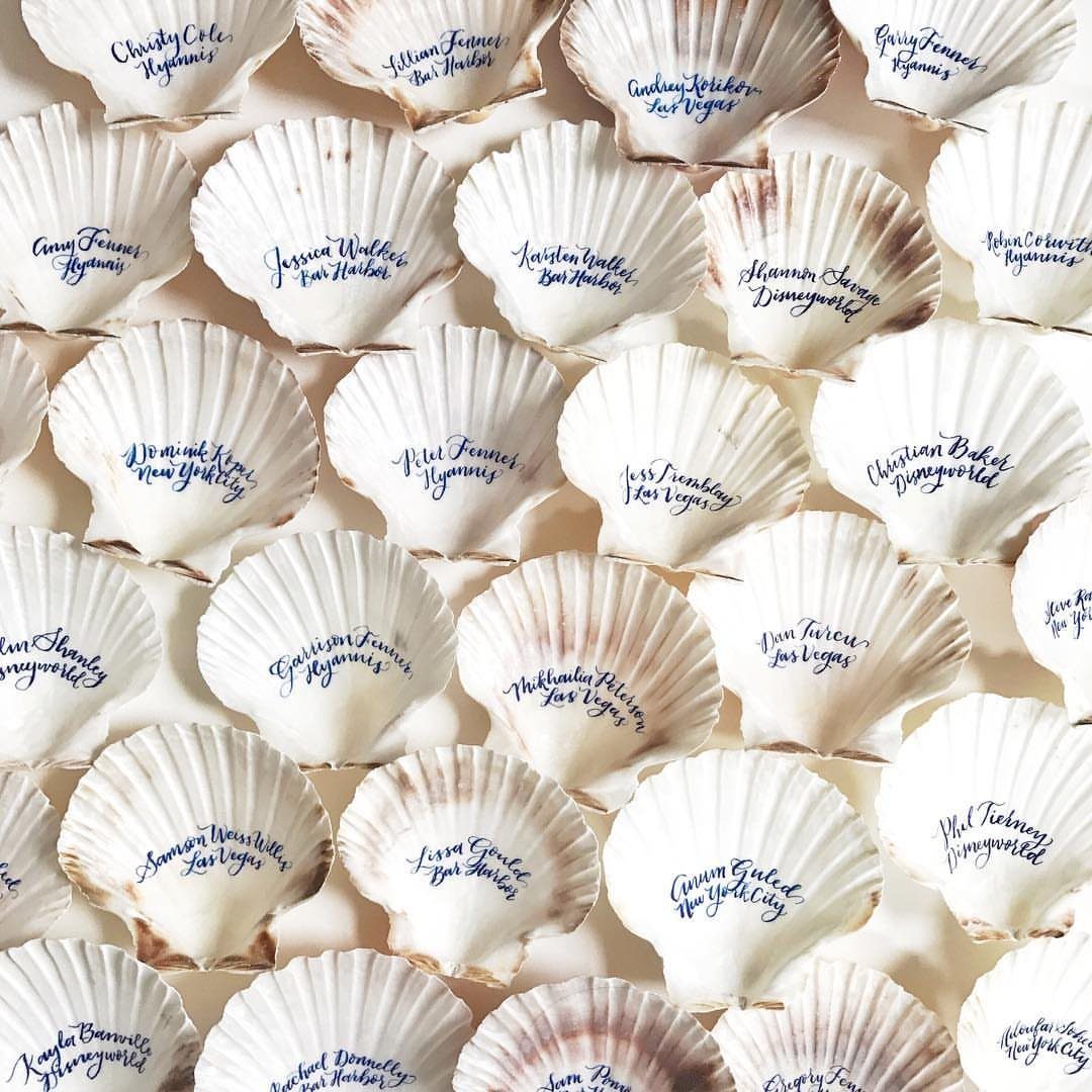 White scallop seashells with handwritten names and locations in blue ink as place cards.