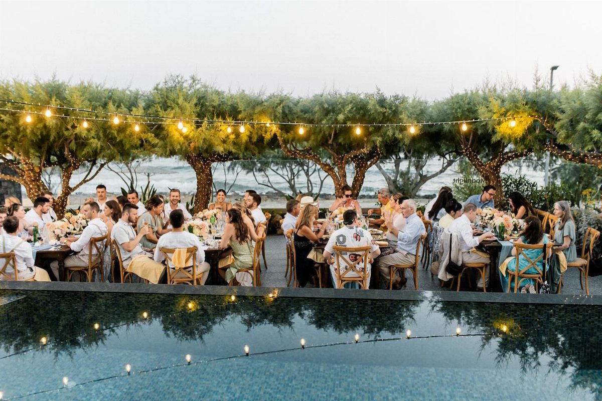 Outdoor wedding dinner with guests at tables under warm hanging string lights by ocean