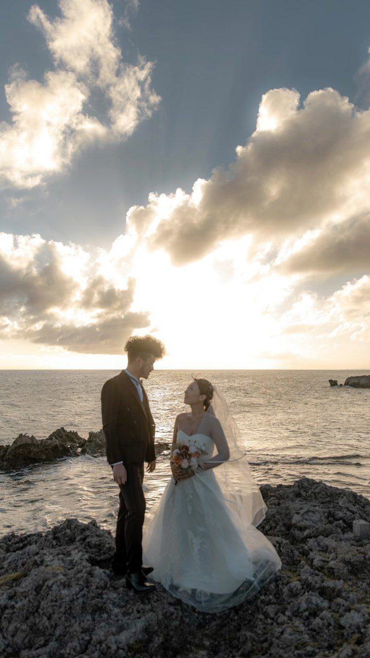 Bride and groom facing each other on a rocky coast during a sunset wedding.