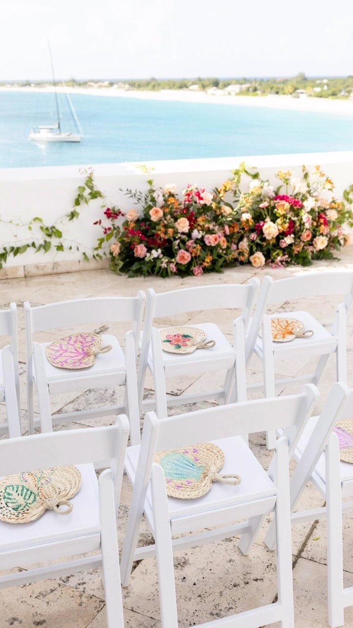 White wedding chairs with colorful woven fans overlooking a turquoise ocean and flowers.