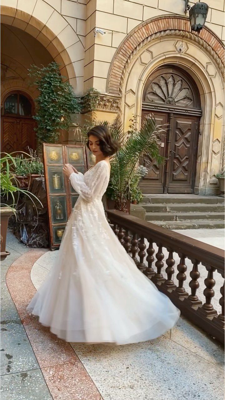 Woman in an elegant long-sleeved white wedding dress standing by a stone archway.