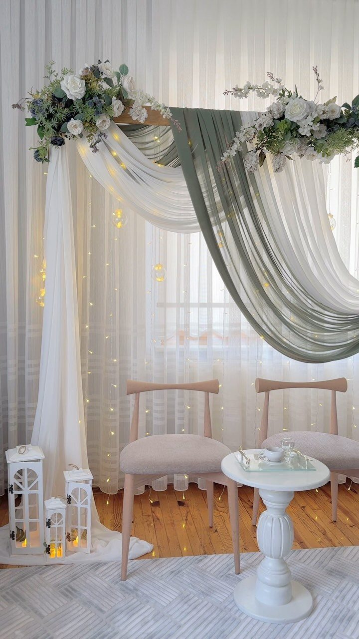 Wedding arch with white and sage green fabric, white flowers, and two chairs.
