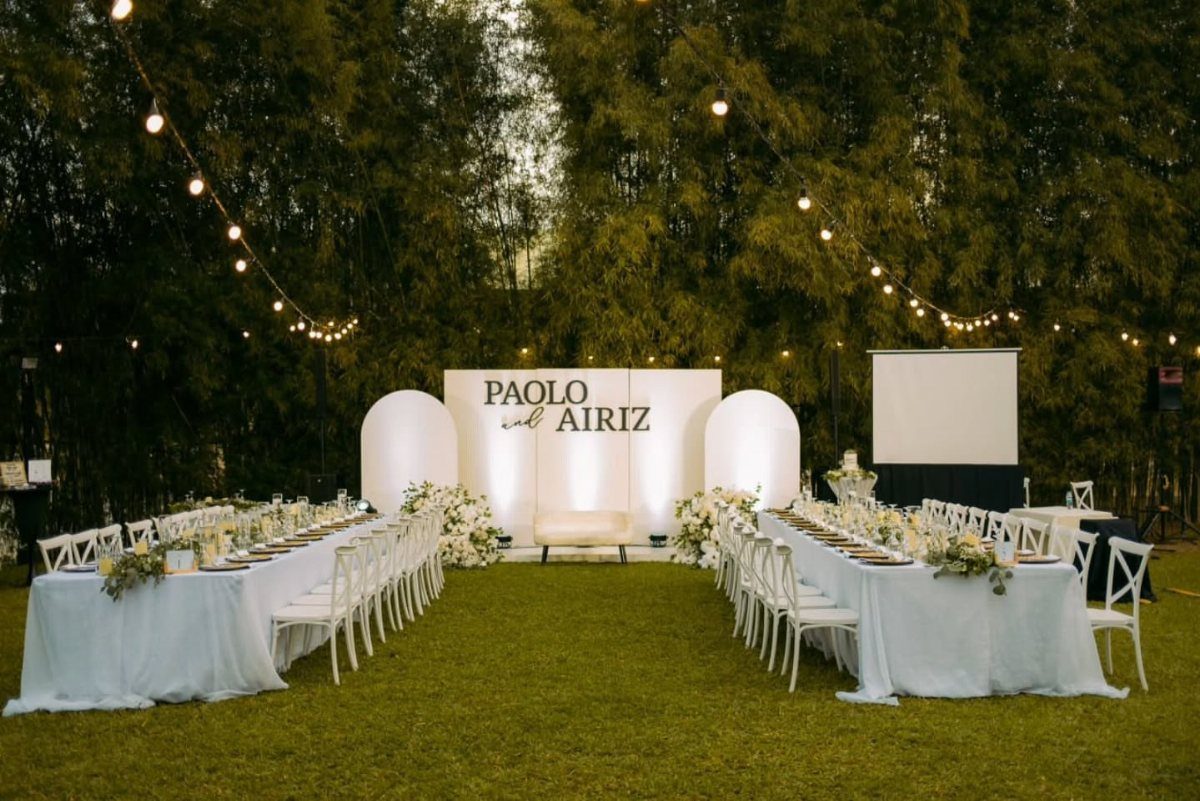 Outdoor wedding reception with long white tables, white chairs, and overhead string lights.