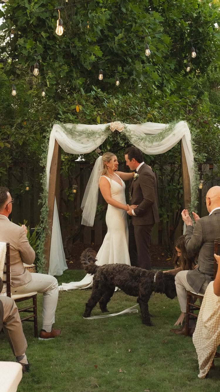 Bride and groom standing under a floral arch during a backyard wedding ceremony.
