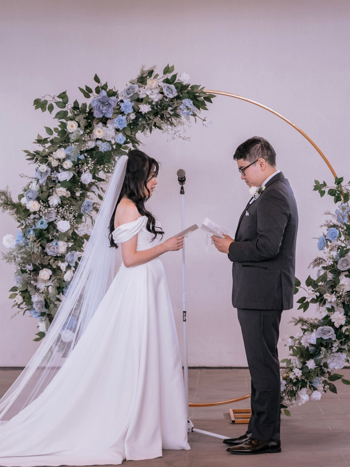 Bride and groom standing and reading vows before a floral circular gold arch.