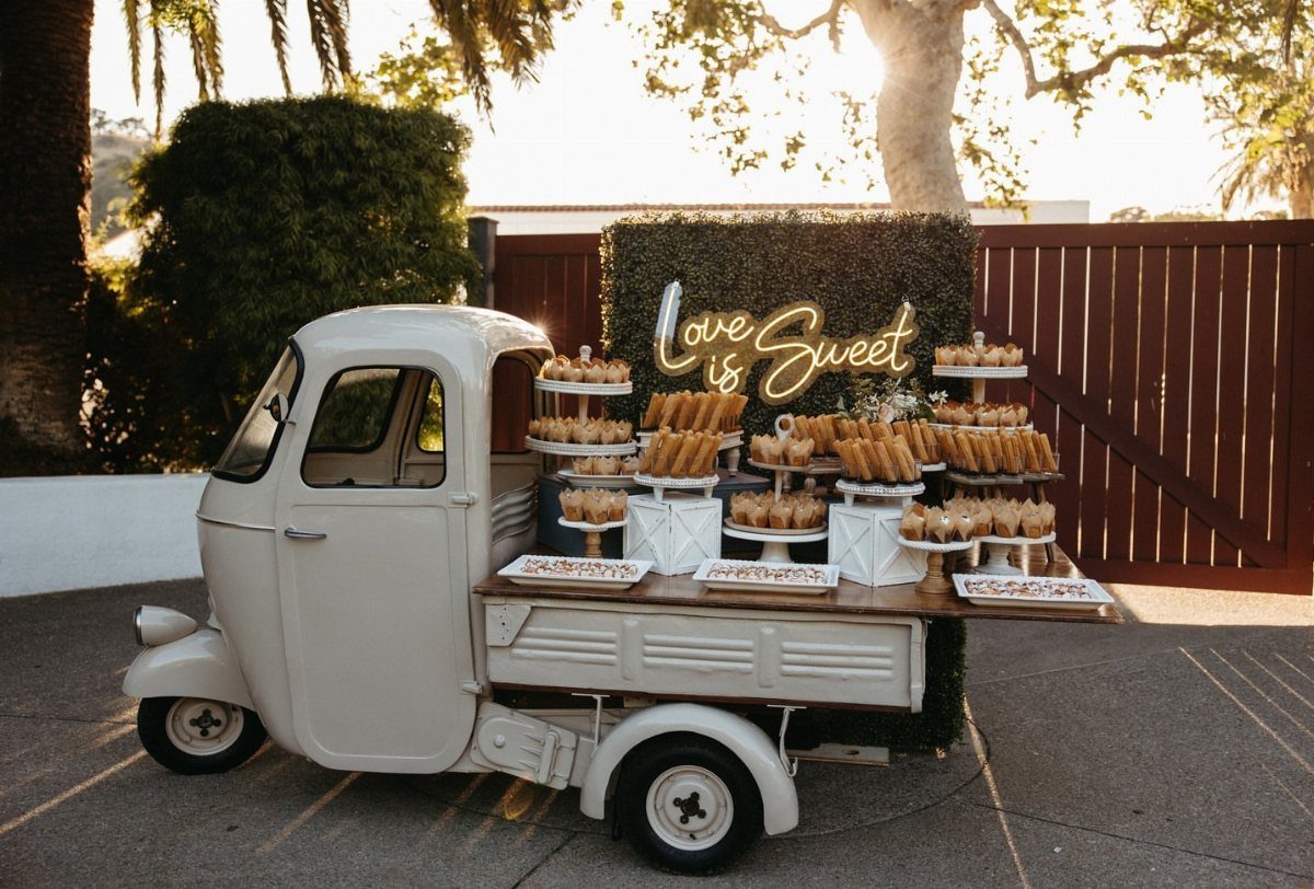 White vintage dessert truck with cupcakes and a Love is Sweet neon sign.
