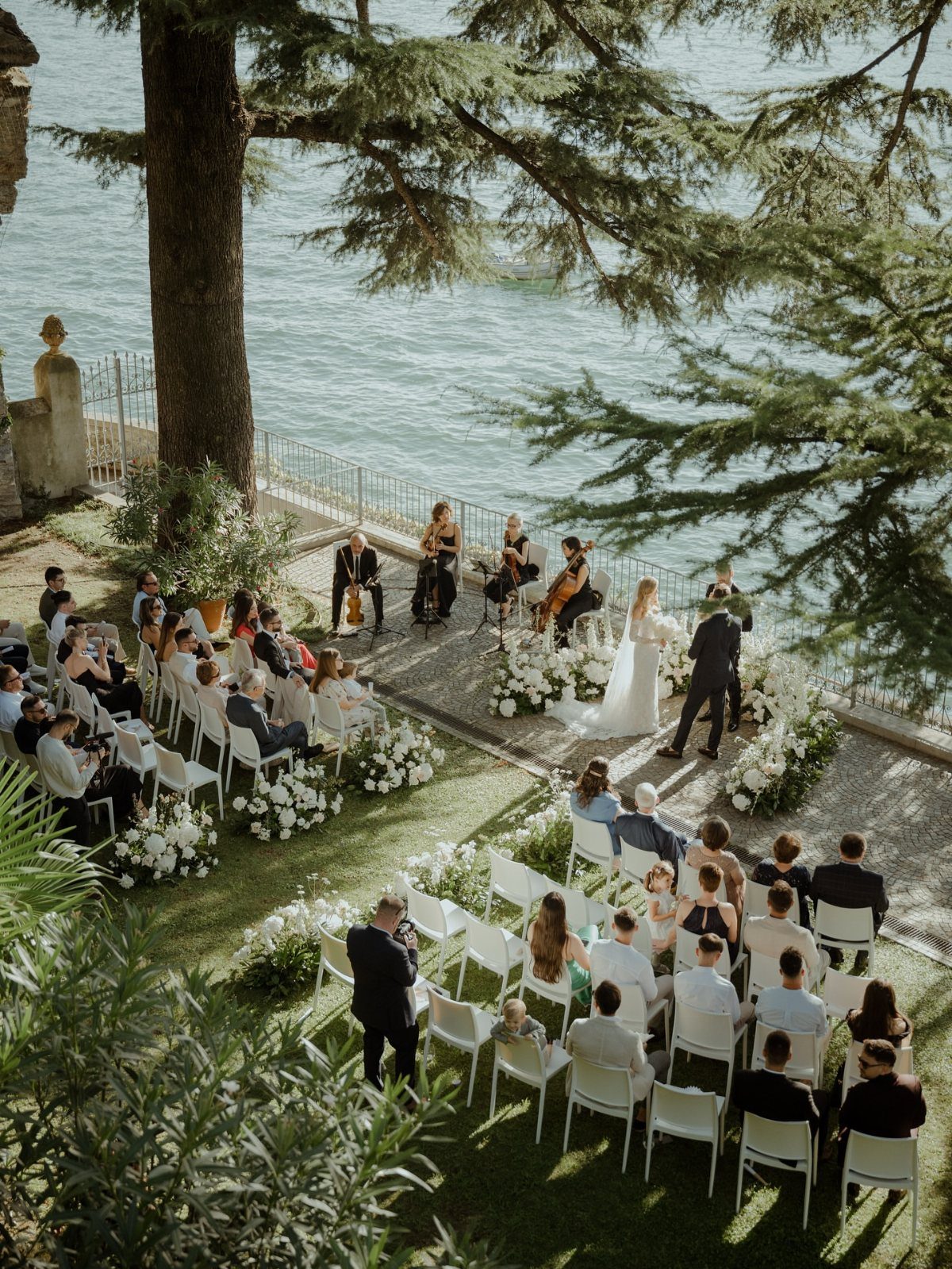 Wedding ceremony by the water with guests seated on a green lawn.