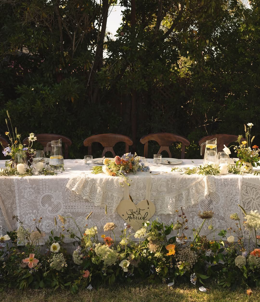 Lace-covered wedding table with floral arrangements set outdoors against a backdrop of trees.