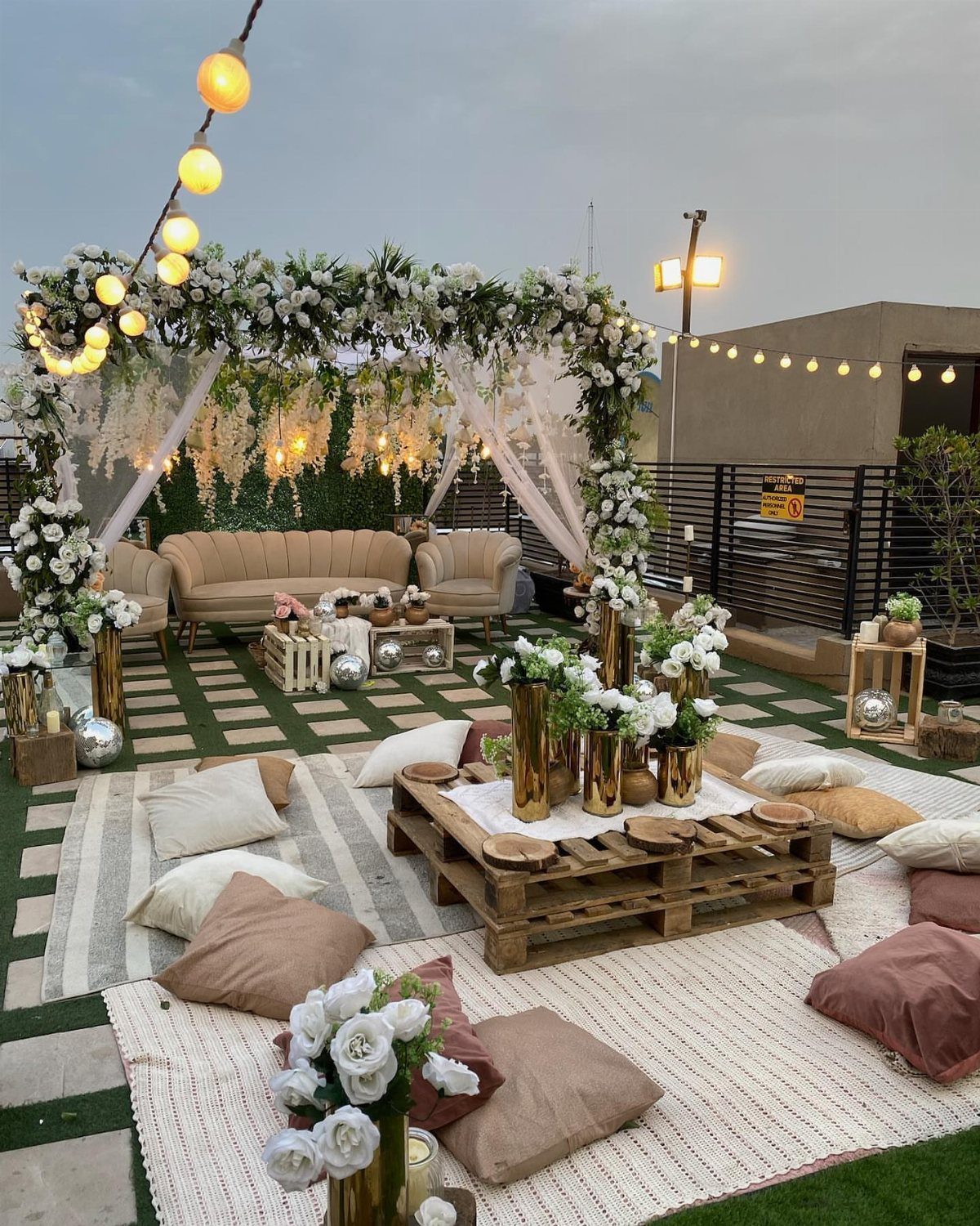 Rooftop lounge with white floral arch, beige seating, and floor cushions on turf.