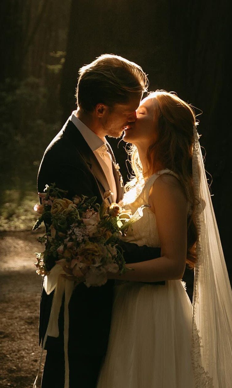 Bride and groom kissing in a sunlit forest, bride holding a floral bouquet.