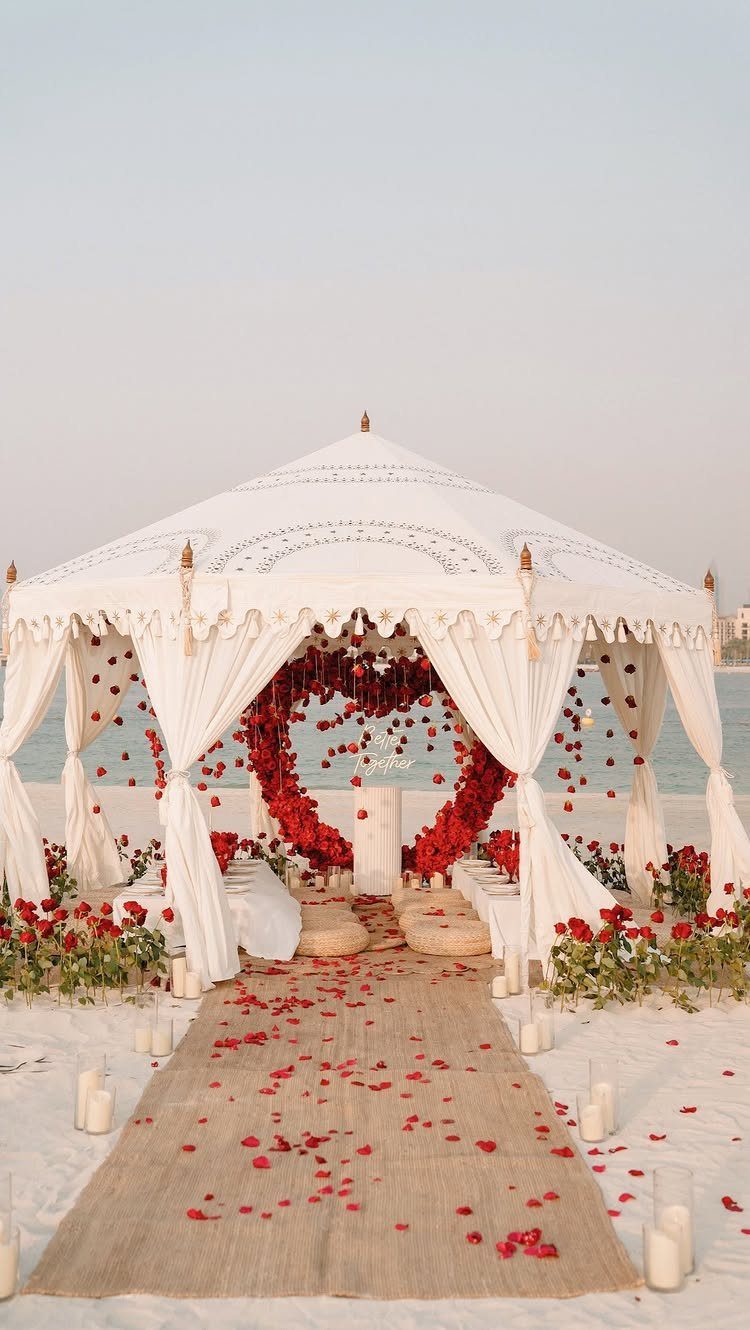 White wedding canopy on a beach with a red rose heart backdrop and candles.