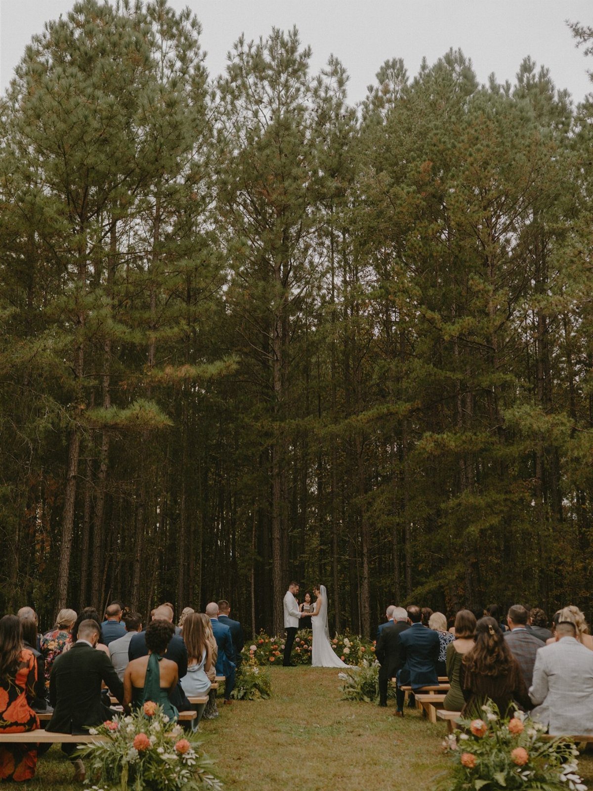 Bride and groom during an outdoor wedding ceremony in a pine forest with guests.