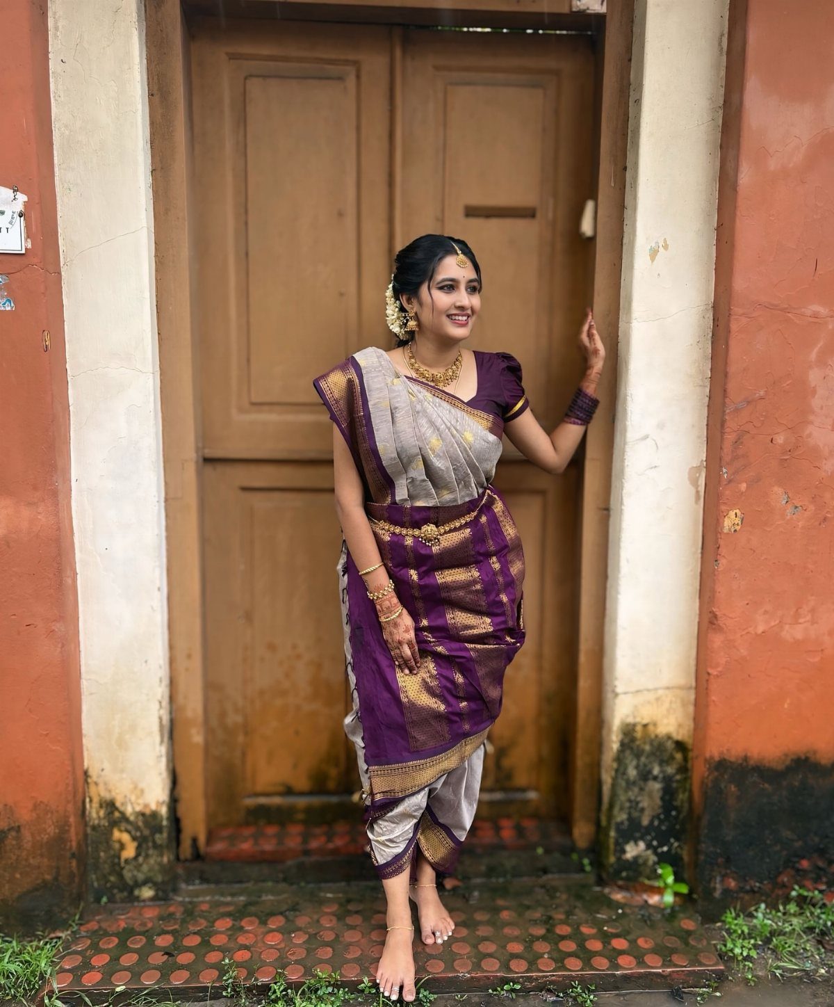 South Indian bride in purple silk saree and gold jewelry standing by a door