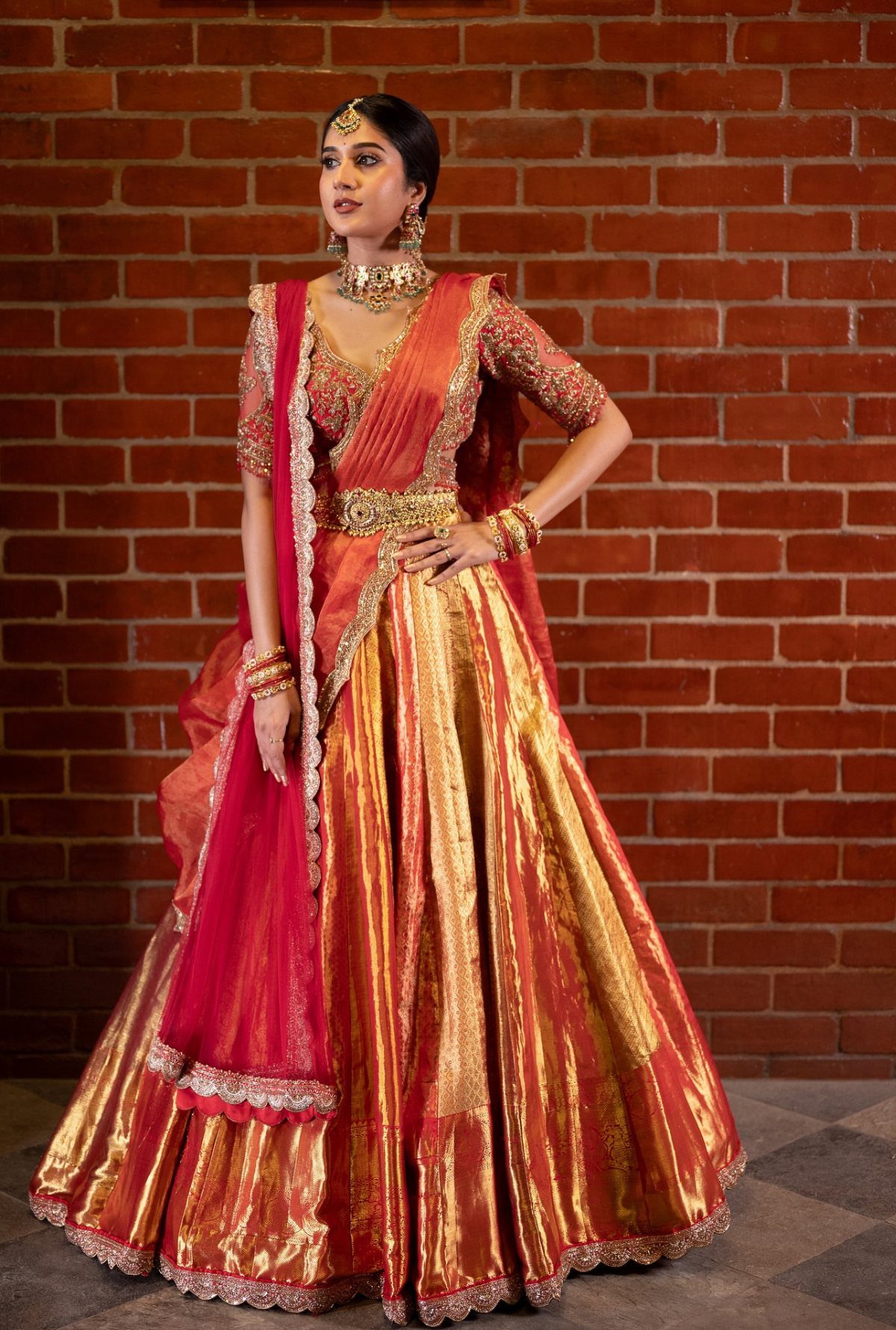 Woman in a red and gold bridal lehenga posing against a brick wall.