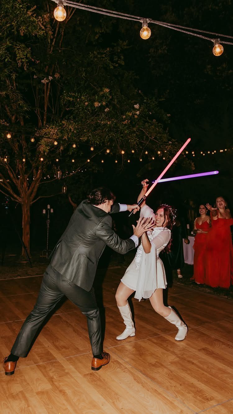 Couple in wedding attire dueling with pink and purple lightsabers on a dance floor.