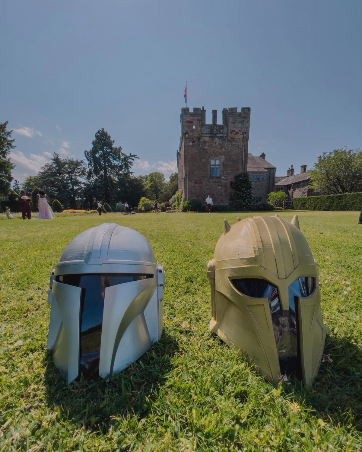 Two Mandalorian helmets on grass with a stone castle in the background.