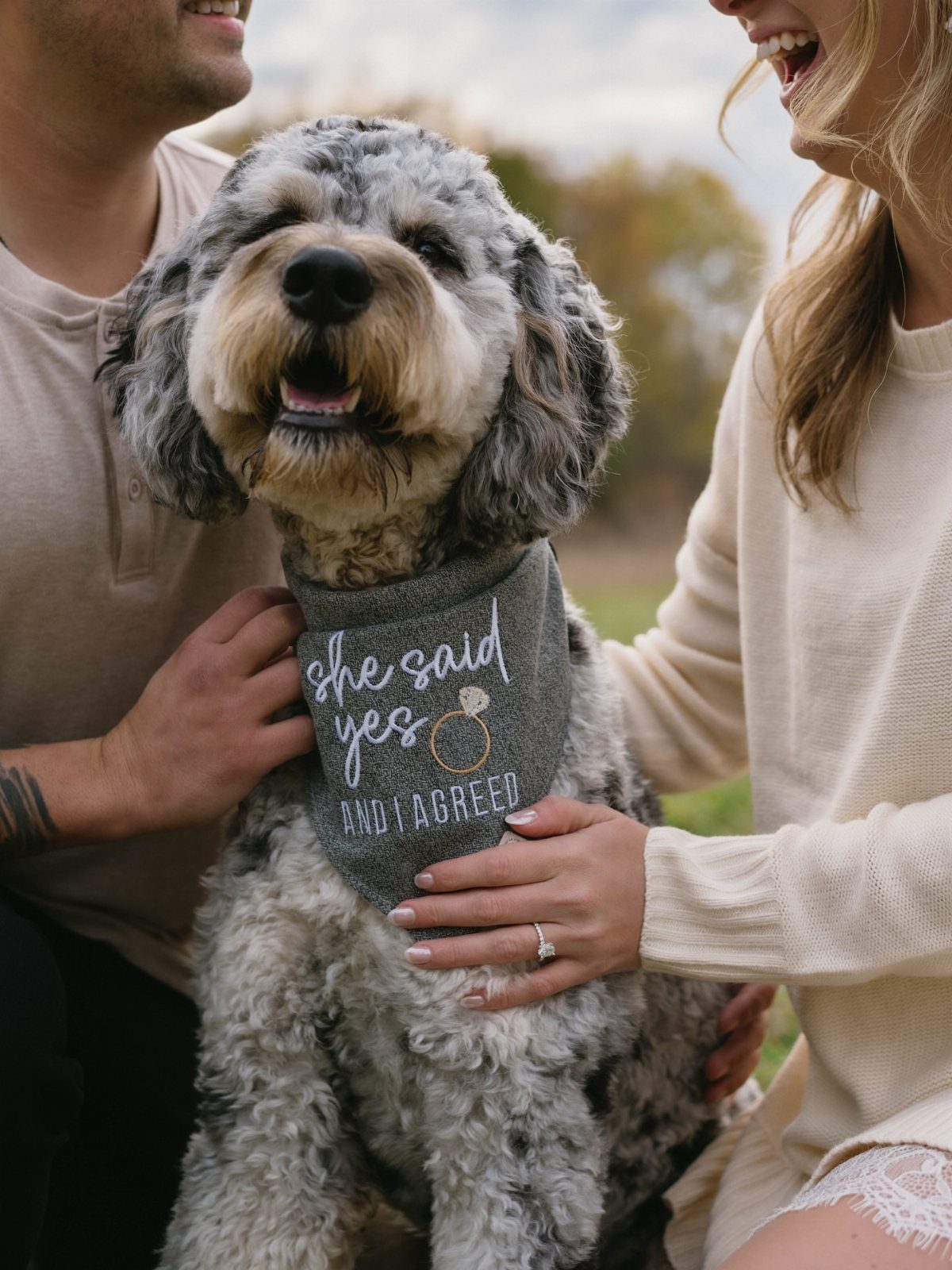 Curly-haired dog wearing an engagement announcement bandana between a smiling couple outdoors.