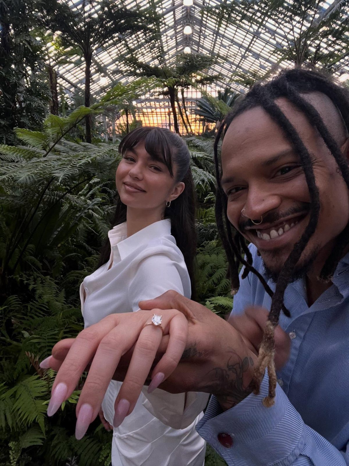 Smiling couple in a greenhouse showing off a diamond engagement ring on a finger