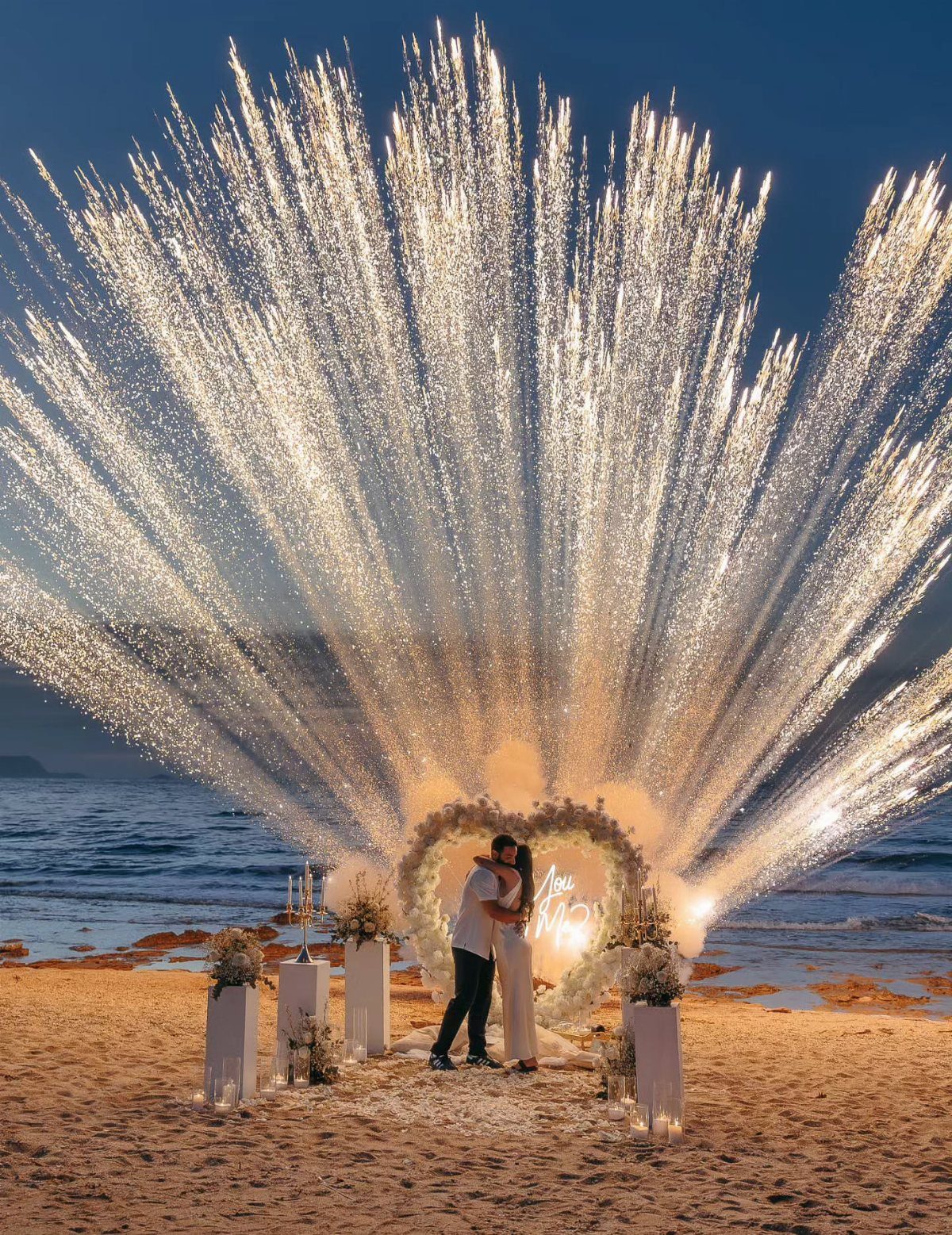 Couple kissing on a beach framed by a floral arch and gold sparklers.