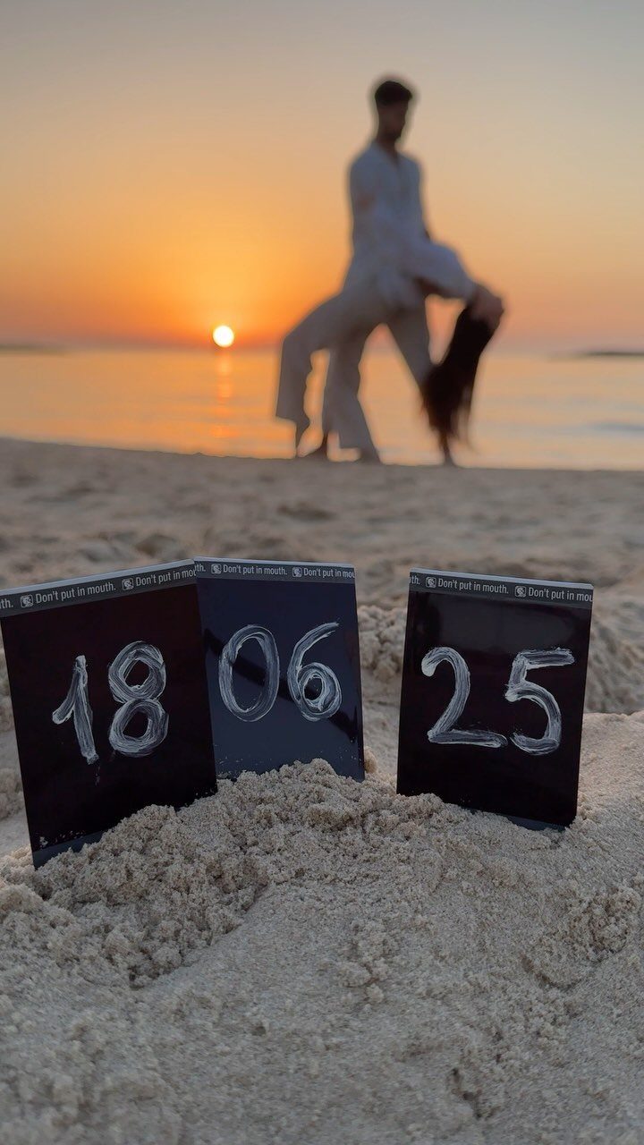 Date cards in beach sand with a blurred couple at sunset behind them.