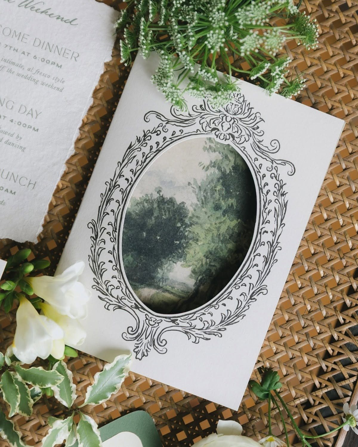 Wedding invitation with an ornate oval frame and landscape painting on wicker surface