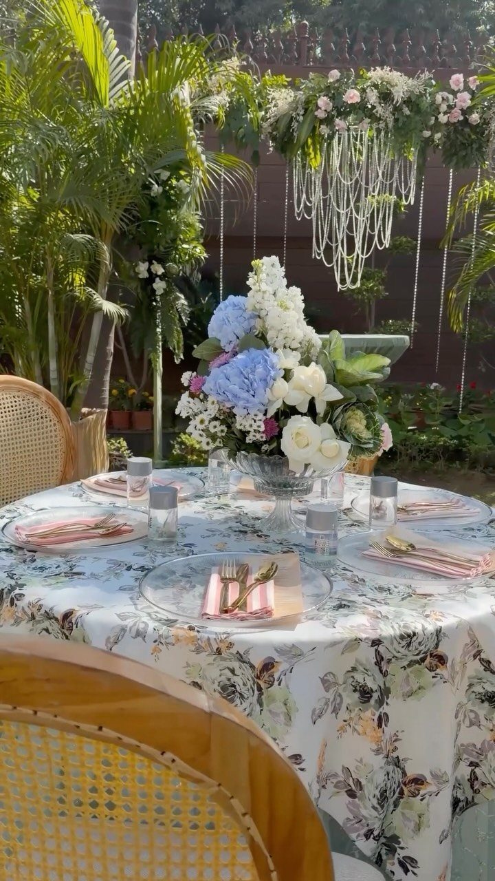 Round wedding table with floral tablecloth, blue flower centerpiece, and wicker chairs outdoors.