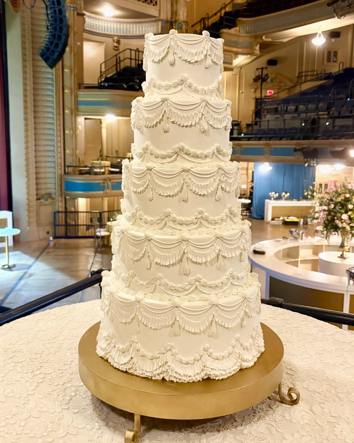 Five-tier white wedding cake with ornate frosting on a gold stand in a theater.