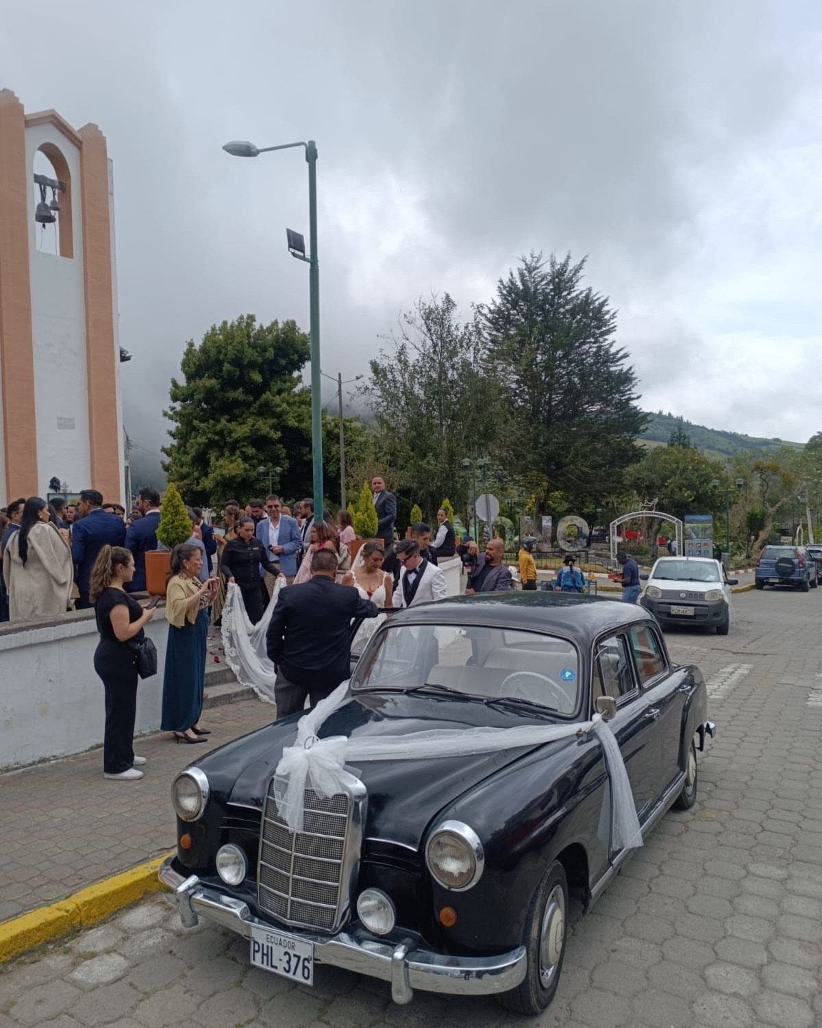 Black vintage car decorated with white wedding ribbons parked near wedding guests.