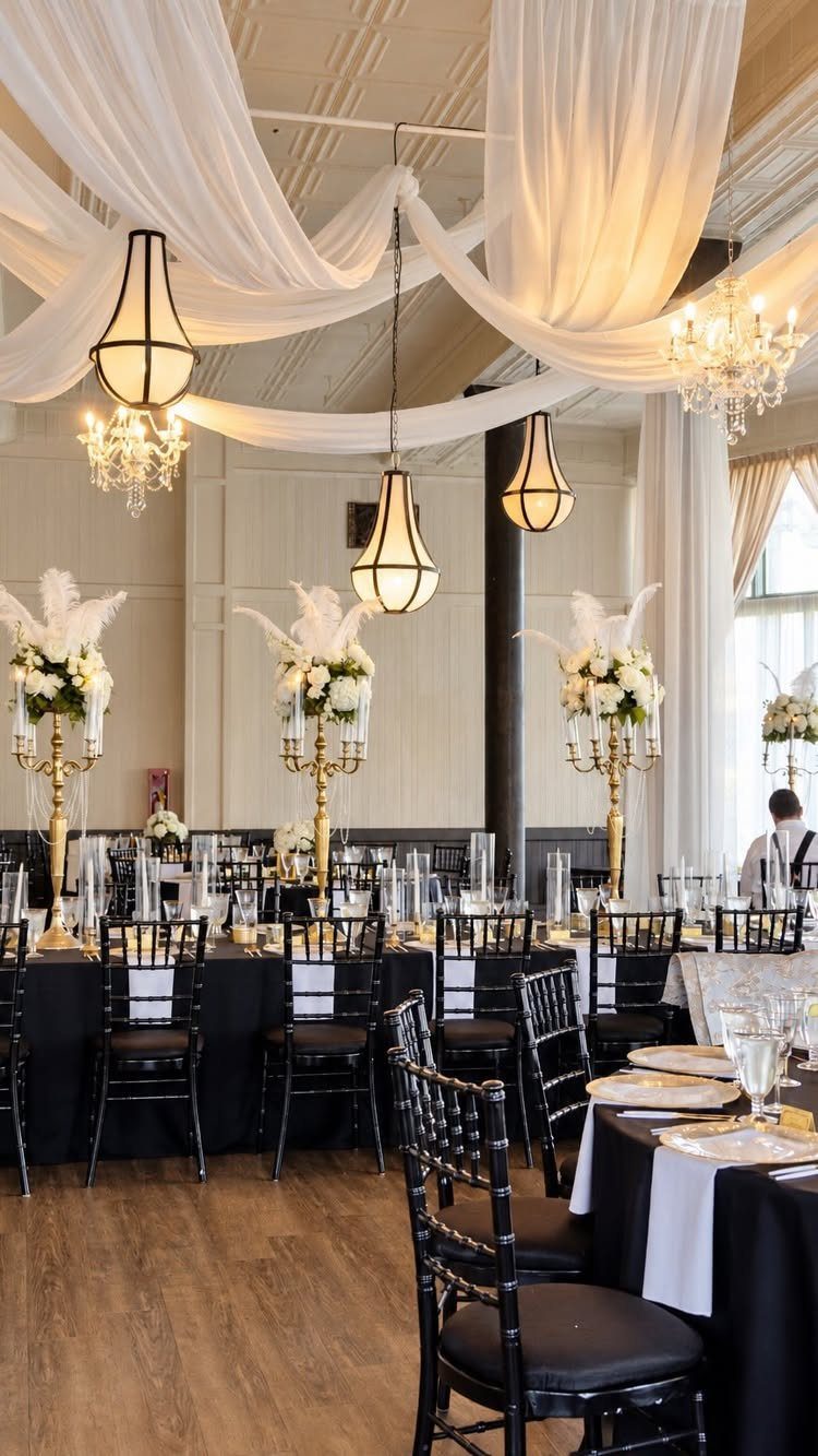 Elegant wedding reception venue with white ceiling drapes, chandeliers, and black table settings.